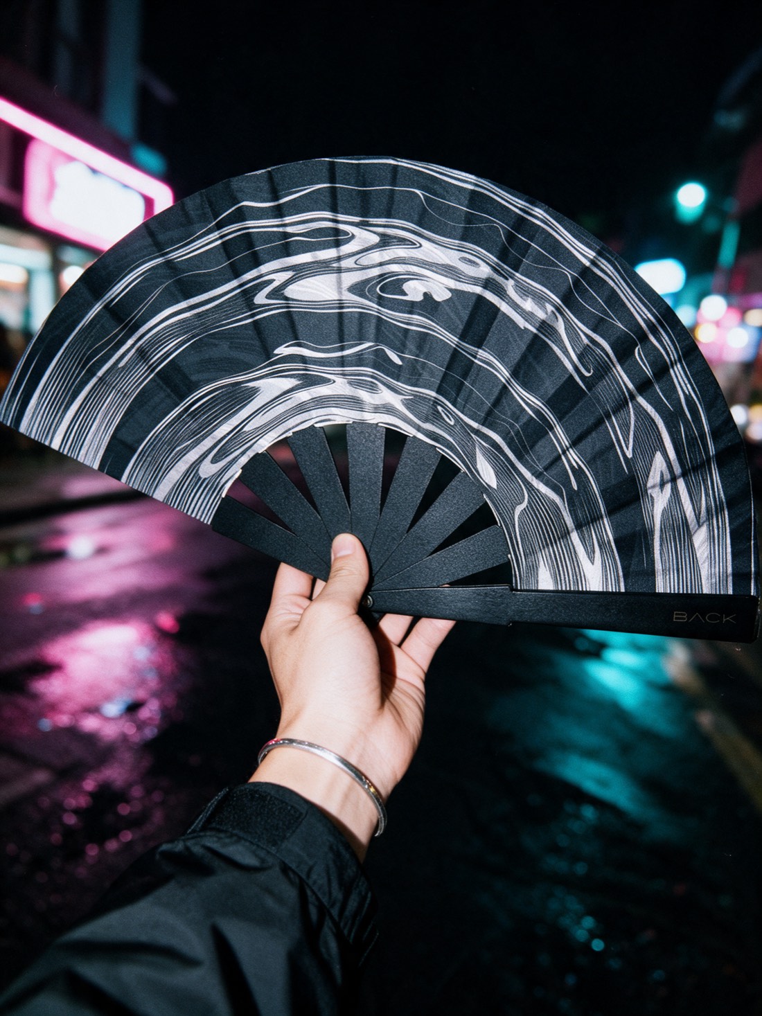 Una mano sostiene hacia la cámara un abanico BACK Rheo en una calle de noche, con luces de neón